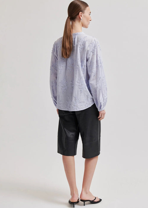 A model wearing a light lavender long-sleeve blouse with textured floral embroidery and a button-up front, paired with black leather pants