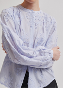 A model wearing a light lavender long-sleeve blouse with textured floral embroidery and a button-up front, paired with black leather pants