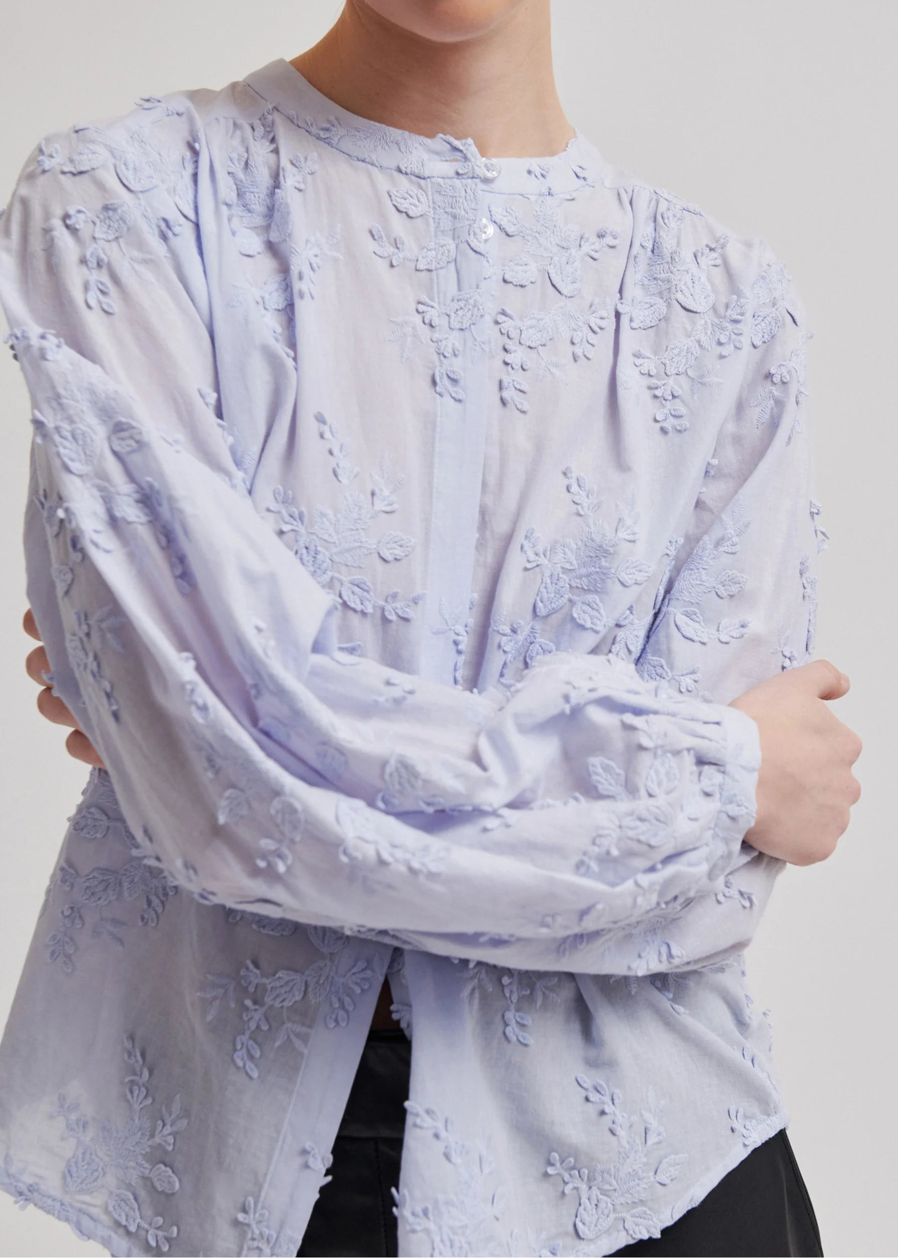 A model wearing a light lavender long-sleeve blouse with textured floral embroidery and a button-up front, paired with black leather pants