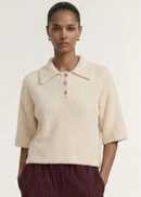 A model wearing a cream-colored, short-sleeved fuzzy knit polo shirt with three buttons, paired with dark burgundy pinstriped trousers, against a plain off-white background.