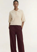 A model wearing a cream-colored, short-sleeved fuzzy knit polo shirt with three buttons, paired with dark burgundy pinstriped trousers, against a plain off-white background.