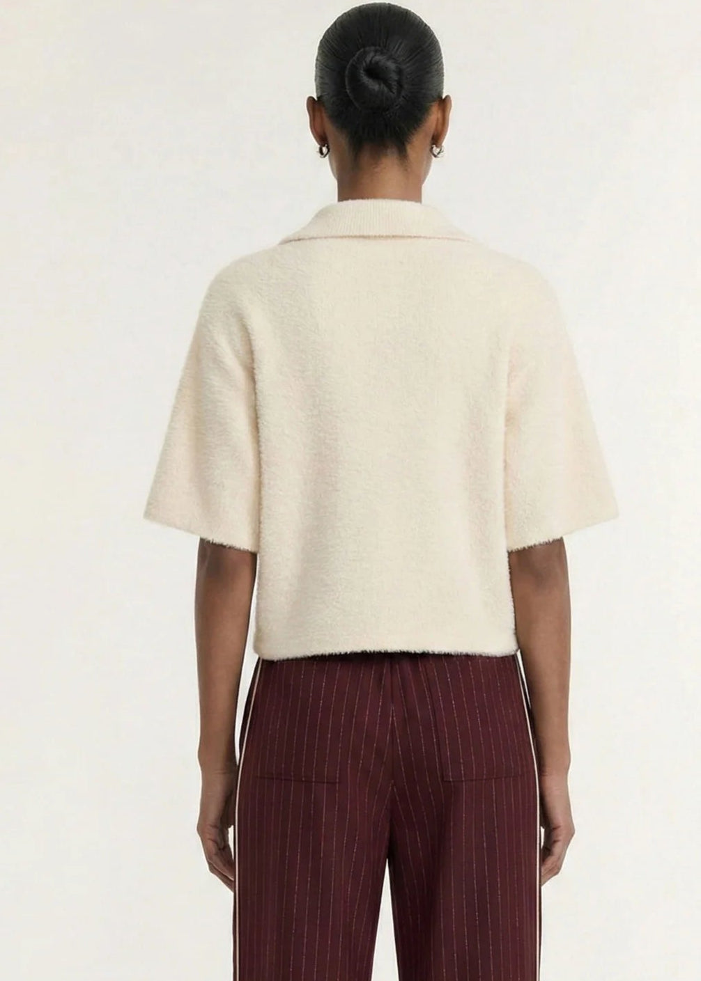 A model wearing a cream-colored, short-sleeved fuzzy knit polo shirt with three buttons, paired with dark burgundy pinstriped trousers, against a plain off-white background.