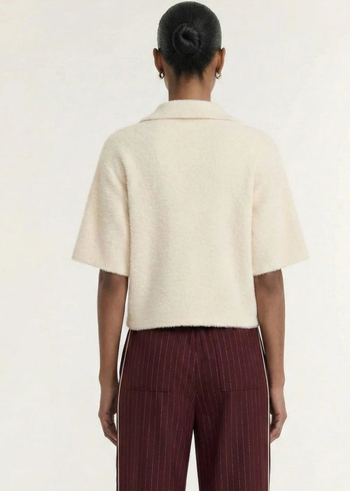 A model wearing a cream-colored, short-sleeved fuzzy knit polo shirt with three buttons, paired with dark burgundy pinstriped trousers, against a plain off-white background.