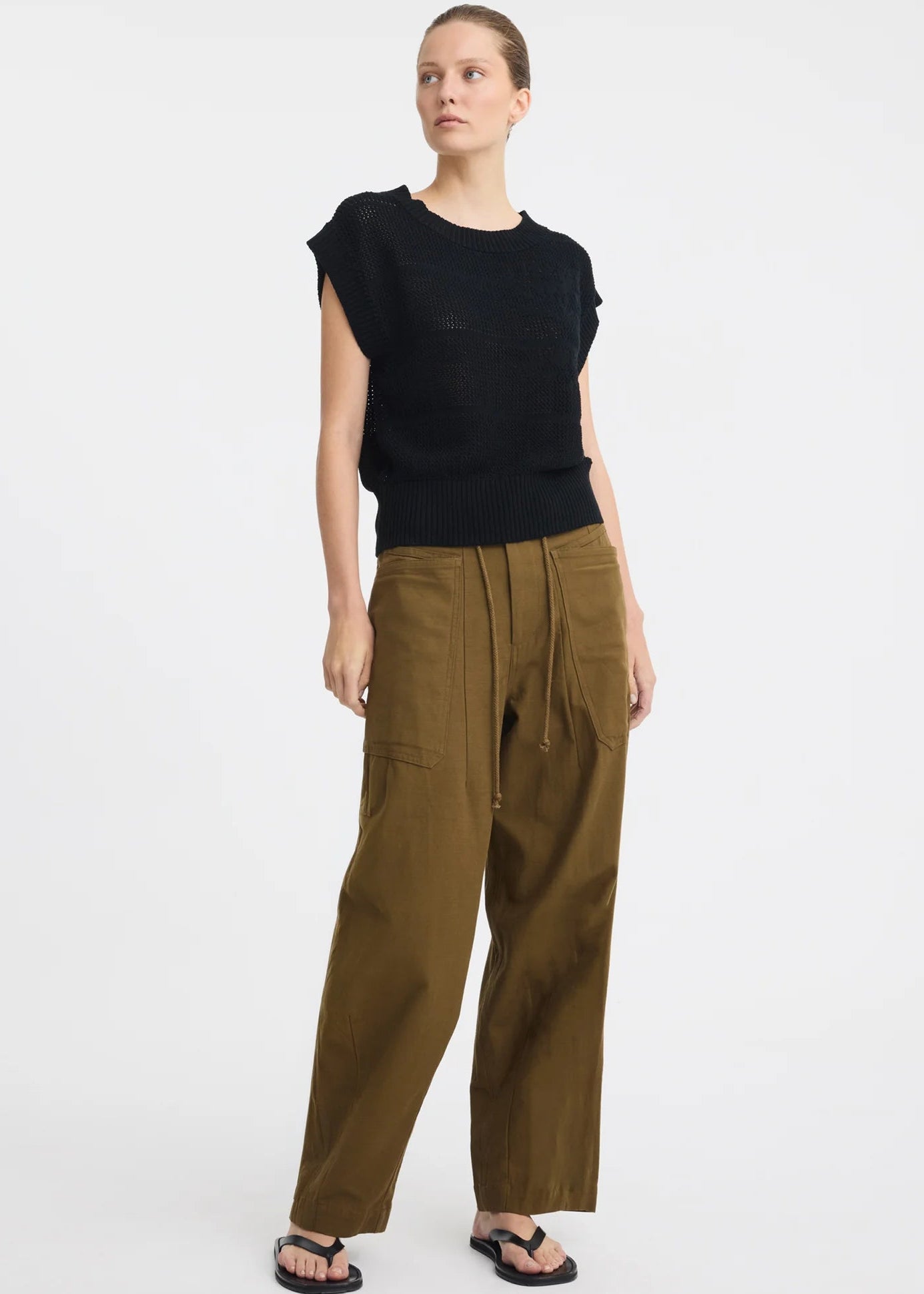 A model wearing a black sleeveless knit top and olive green wide-leg pants with a drawstring waist, paired with black sandals, against a plain white studio background.
