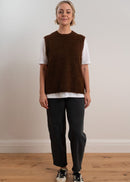 Person wearing a brown knitted vest over a white shirt with dark pants on a plain background