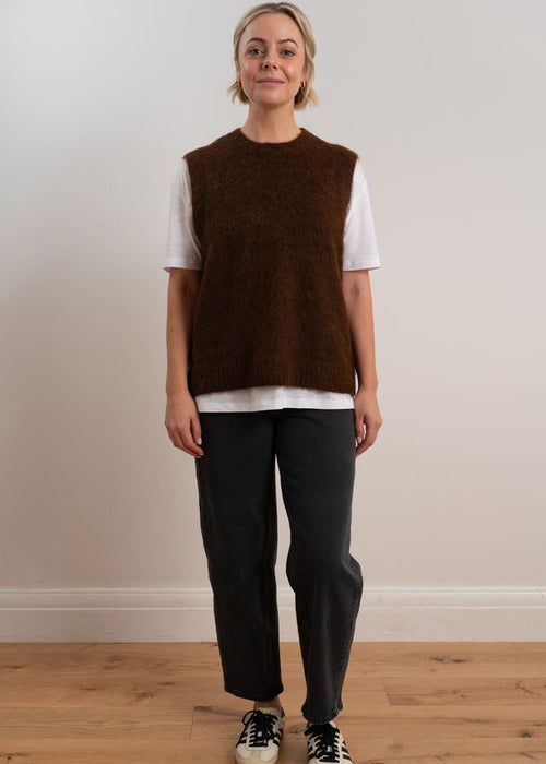Person wearing a brown knitted vest over a white shirt with dark pants on a plain background