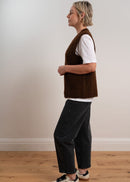 Person wearing a brown knitted vest over a white shirt with dark pants on a plain background