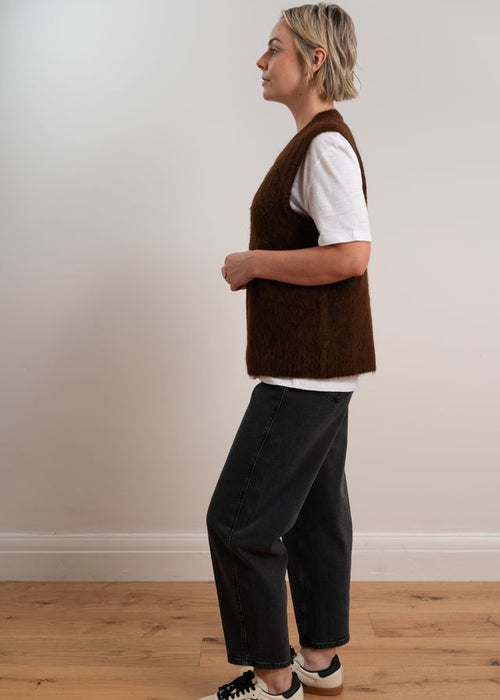 Person wearing a brown knitted vest over a white shirt with dark pants on a plain background