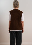 Person wearing a brown knitted vest over a white shirt with dark pants on a plain background