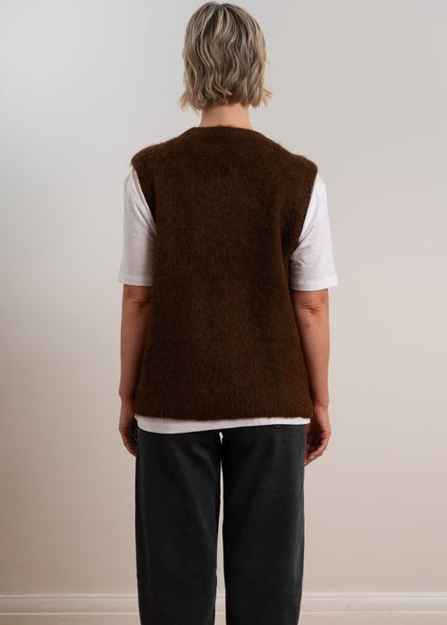 Person wearing a brown knitted vest over a white shirt with dark pants on a plain background