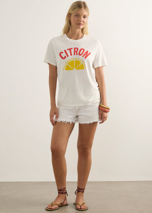 A woman modelling a white graphic t-shirt with a red 'CITRON' text and yellow lemon slice design, paired with white frayed denim shorts and brown sandals, against a plain light grey studio background.