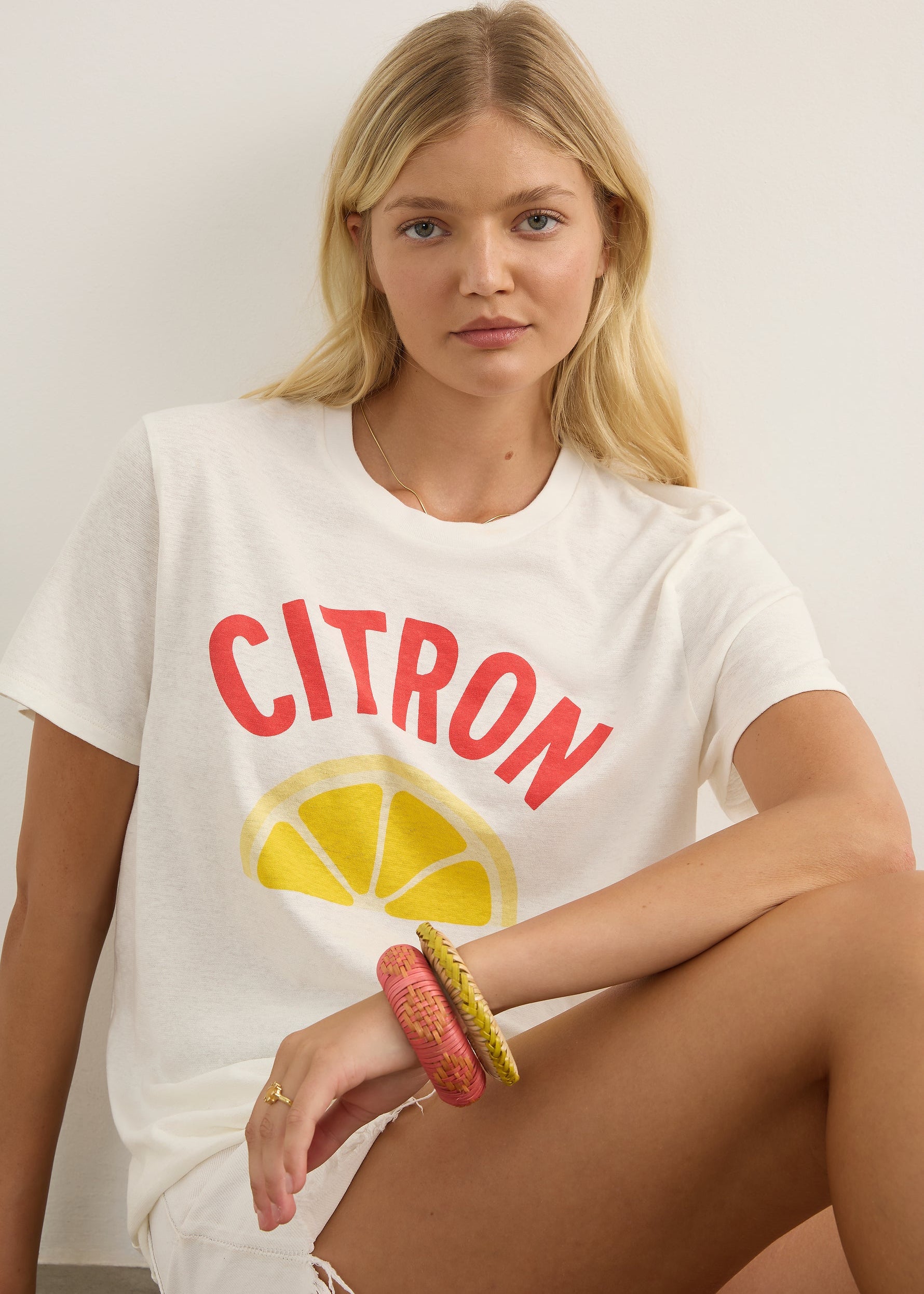 A woman modelling a white graphic t-shirt with a red 'CITRON' text and yellow lemon slice design, paired with white frayed denim shorts and brown sandals, against a plain light grey studio background.