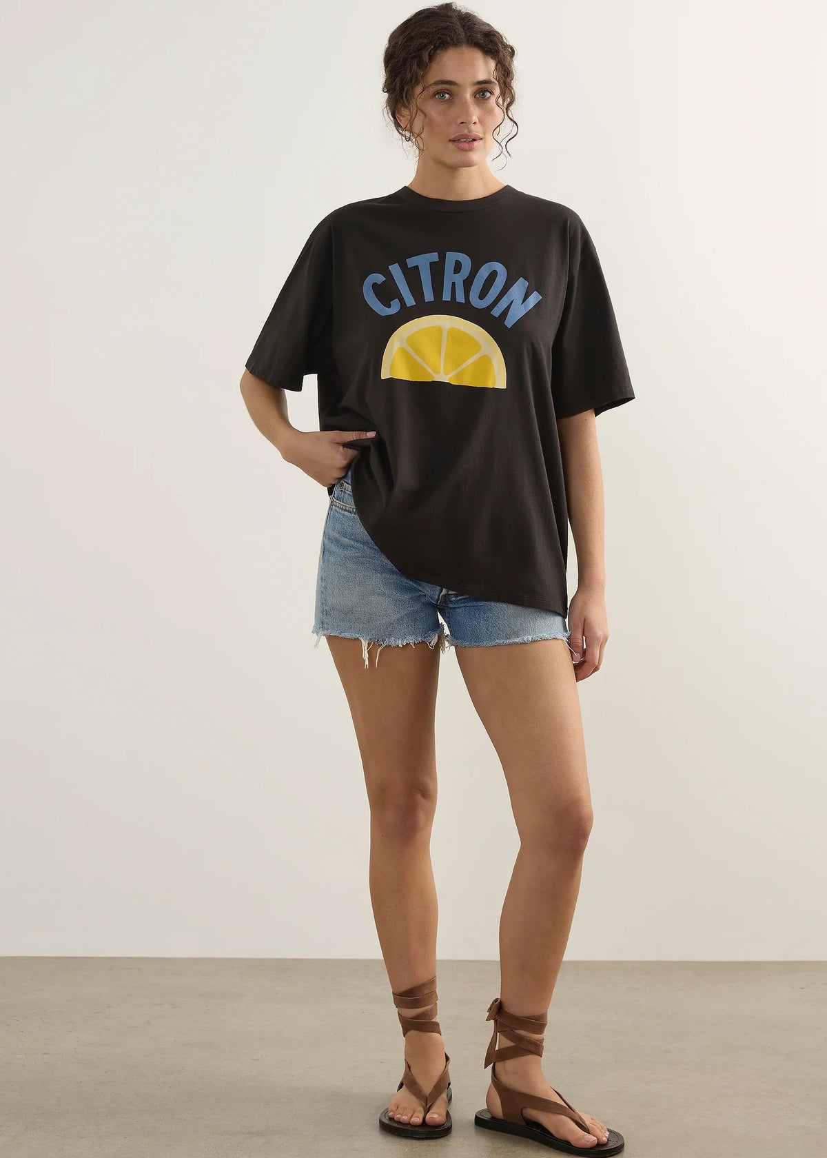 A woman modelling an oversized black t-shirt with a yellow lemon graphic and the word 'CITRON' in blue, paired with denim shorts and brown strappy sandals, set against a minimalist studio background.