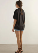 A woman shown from the back wearing an oversized black t-shirt, denim shorts, and brown strappy sandals, standing against a plain off-white wall and light grey floor.