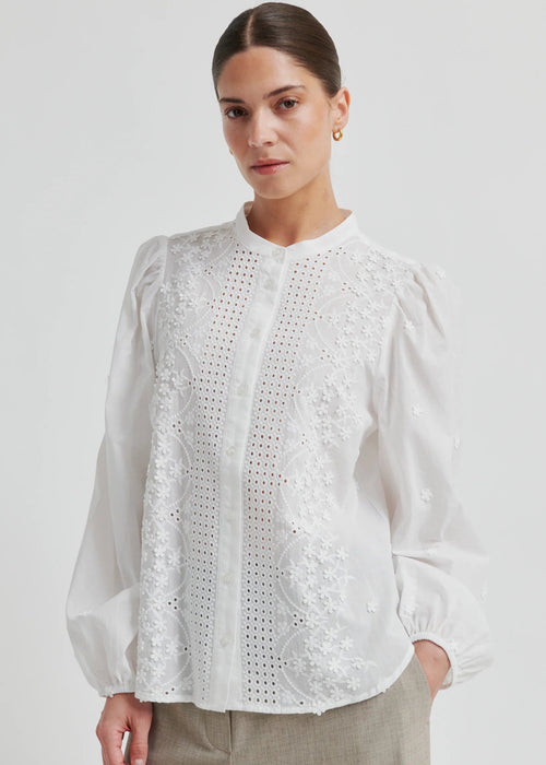 Woman wearing a white blouse with intricate patterns on a plain background
