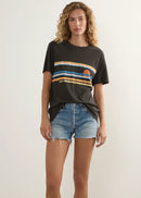 A model stands in a studio wearing a dark charcoal graphic t-shirt with a horizontal striped sunset design, light-wash denim shorts, and white slide sandals.