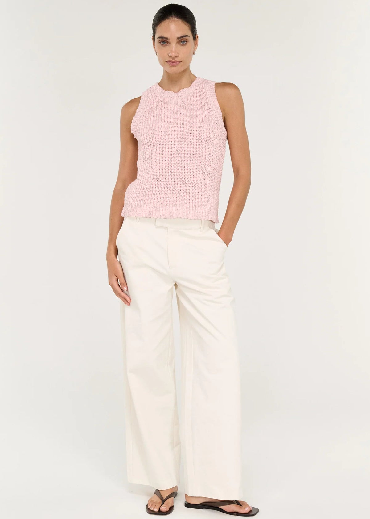 A woman modelling a light pink sleeveless knit top with white trousers, standing against a plain white studio background.
