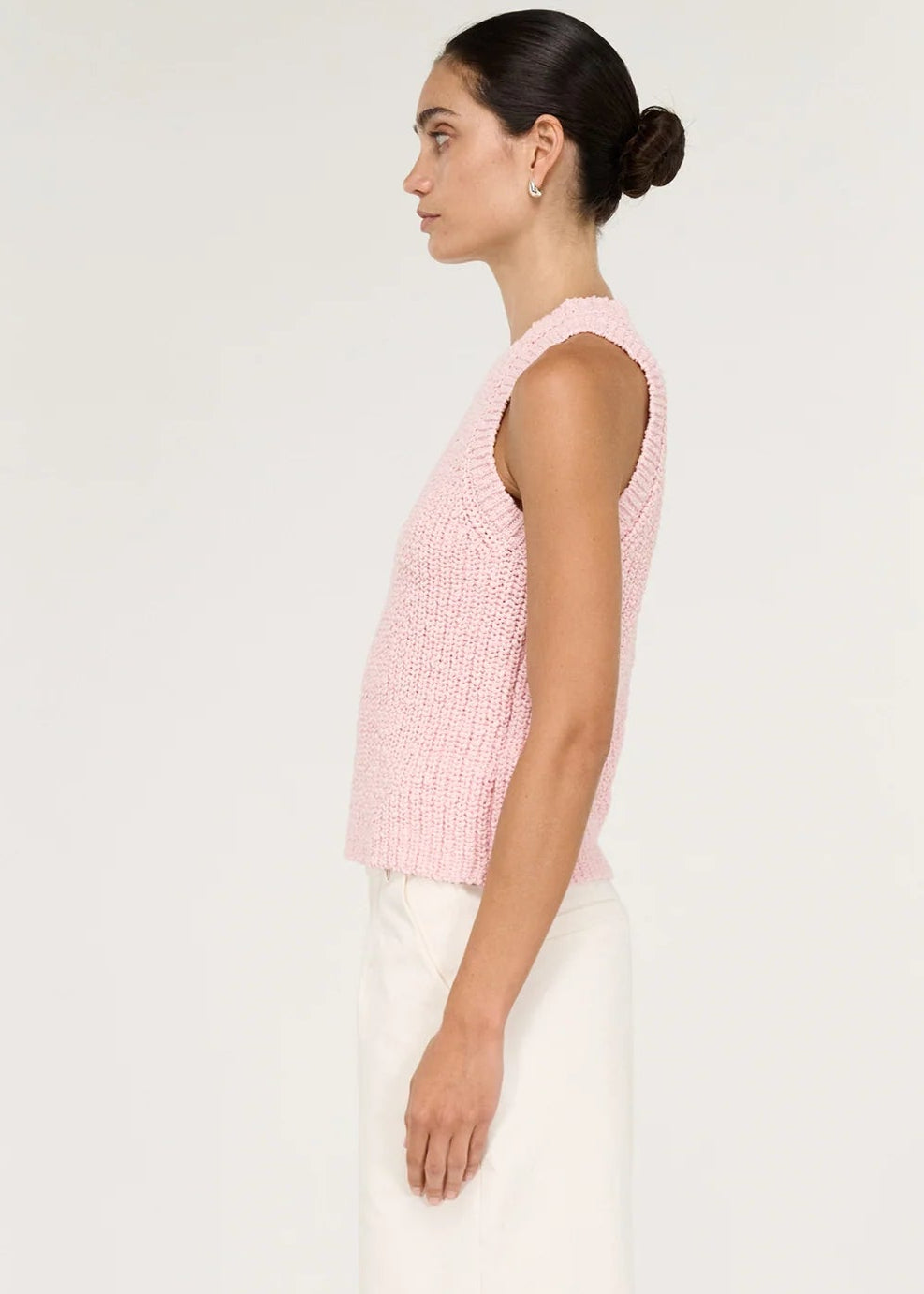 A woman modelling a light pink sleeveless knit top with white trousers, standing against a plain white studio background.