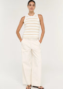 A woman modelling a sleeveless knit top with horizontal beige stripes on a white textured background, paired with white trousers, against a light grey studio backdrop.