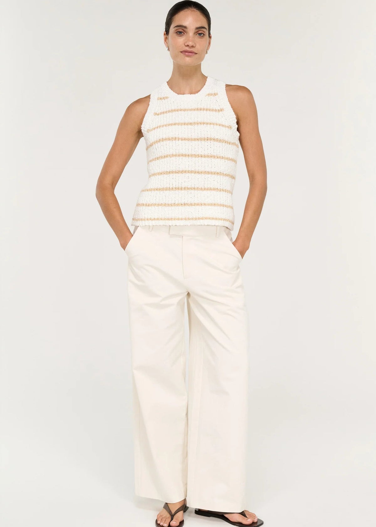 A woman modelling a sleeveless knit top with horizontal beige stripes on a white textured background, paired with white trousers, against a light grey studio backdrop.