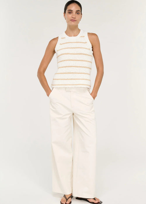 A woman modelling a sleeveless knit top with horizontal beige stripes on a white textured background, paired with white trousers, against a light grey studio backdrop.