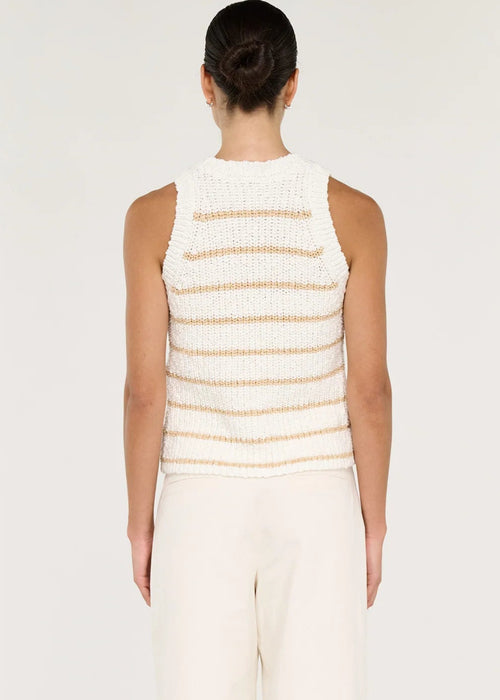 A woman modelling a sleeveless knit top with horizontal beige stripes on a white textured background, paired with white trousers, against a light grey studio backdrop.
