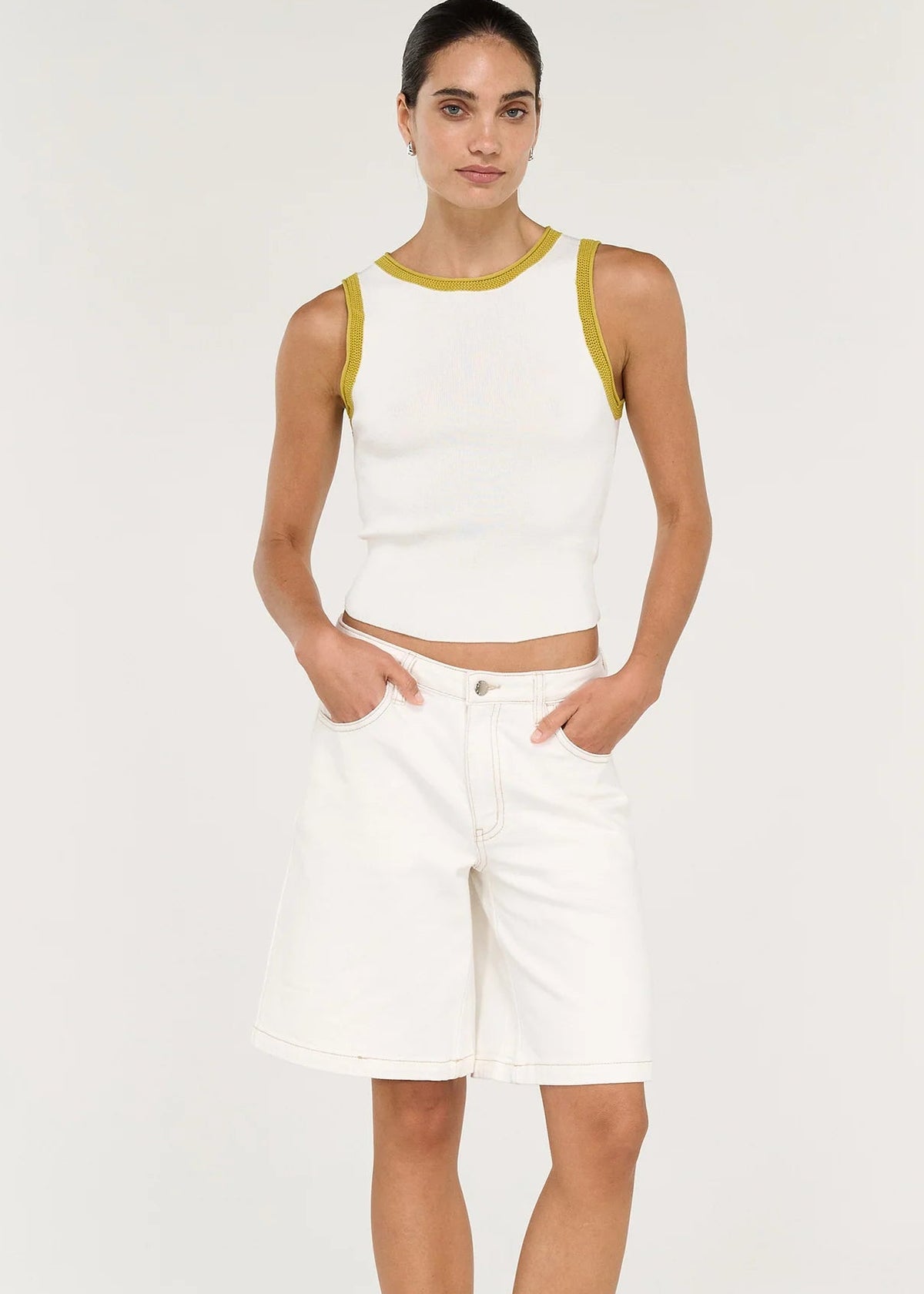 A woman modelling a white ribbed knit sleeveless crop top with yellow trim and white denim shorts against a plain light background.