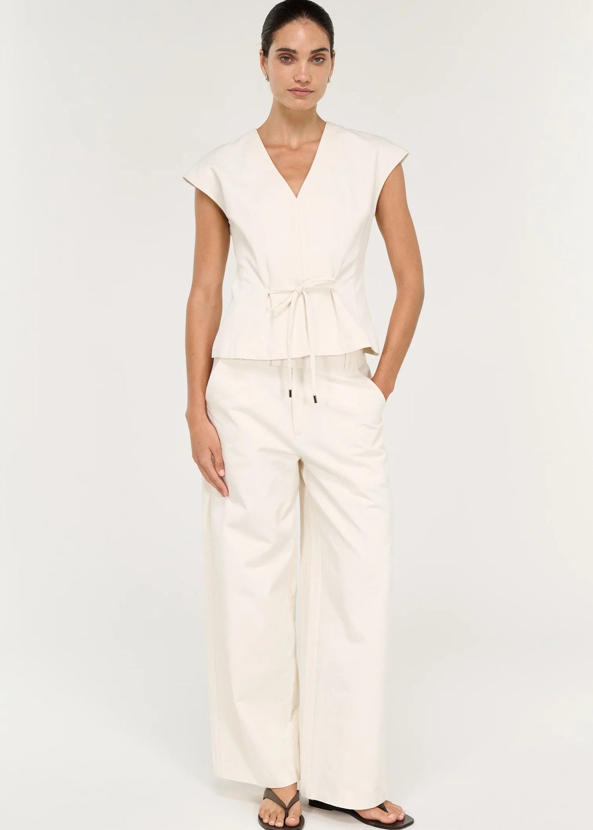 A woman modelling a cream-colored wrap-style top with a tie waist and matching wide-leg pants against a plain white studio background.