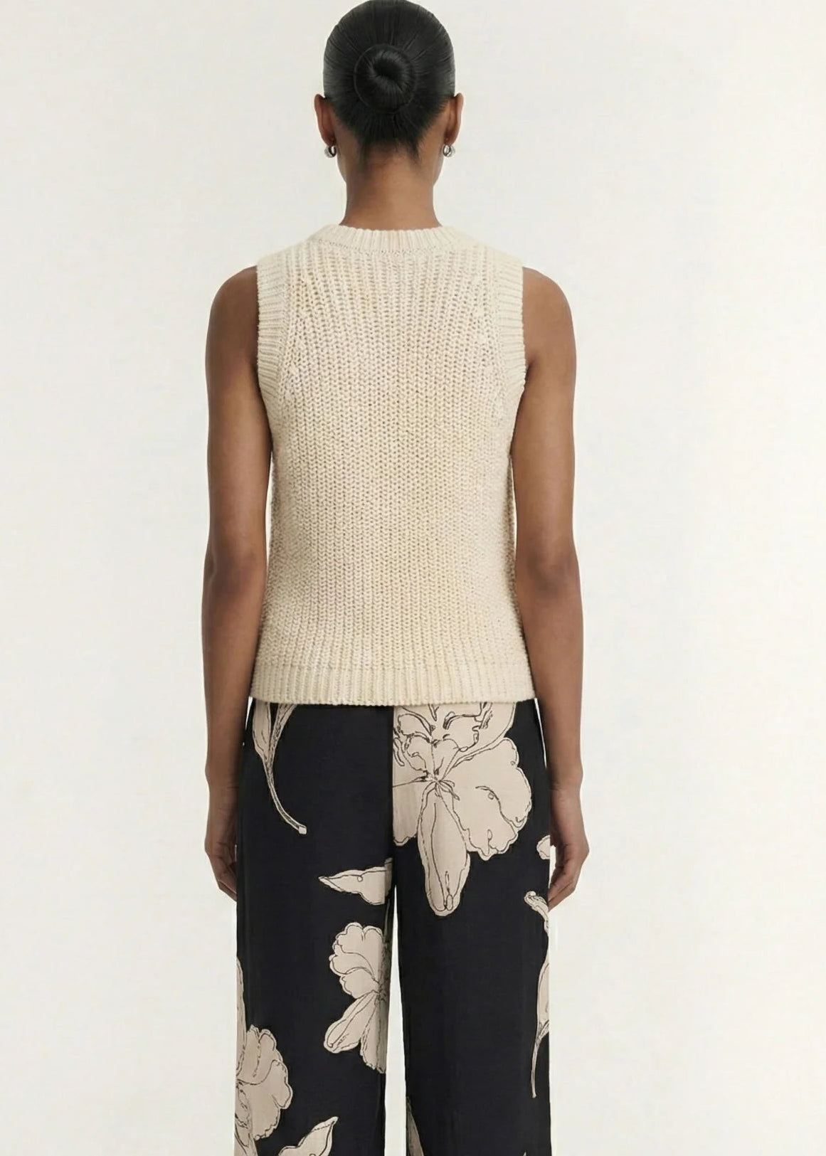 A woman modelling a sleeveless cream-colored ribbed knit top paired with black floral pants against a plain light background.