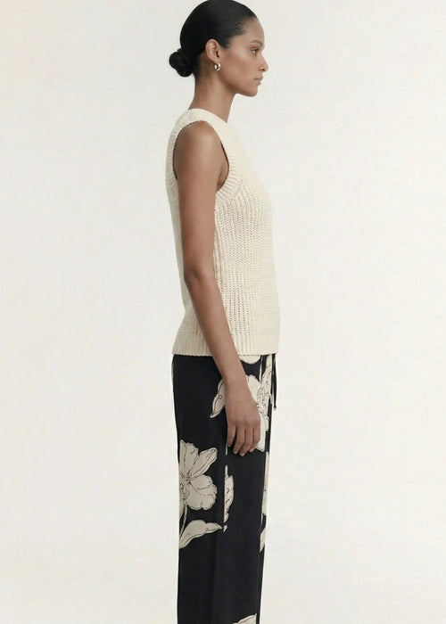 A woman modelling a sleeveless cream-colored ribbed knit top paired with black floral pants against a plain light background.