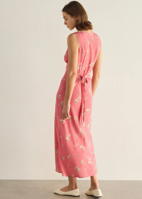 A woman modelling a sleeveless pink midi dress with a delicate white and green floral print, standing against a neutral light grey studio background.