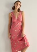 A woman modelling a sleeveless pink midi dress with a delicate white and green floral print, standing against a neutral light grey studio background.