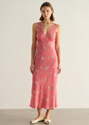 A woman modelling a sleeveless pink midi dress with a delicate white and green floral print, standing against a neutral light grey studio background.