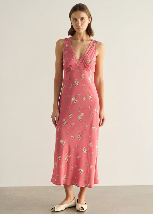 A woman modelling a sleeveless pink midi dress with a delicate white and green floral print, standing against a neutral light grey studio background.