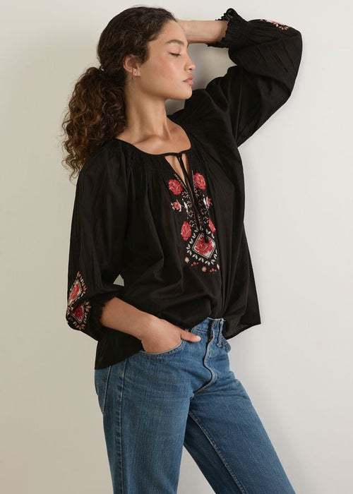 Woman wearing a black blouse with floral embroidery and blue jeans against a plain background