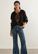 Woman wearing a black blouse with floral embroidery and blue jeans against a plain background
