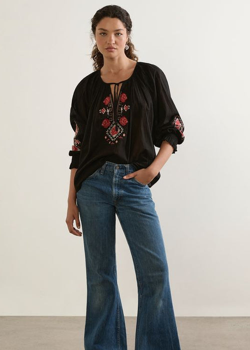 Woman wearing a black blouse with floral embroidery and blue jeans against a plain background