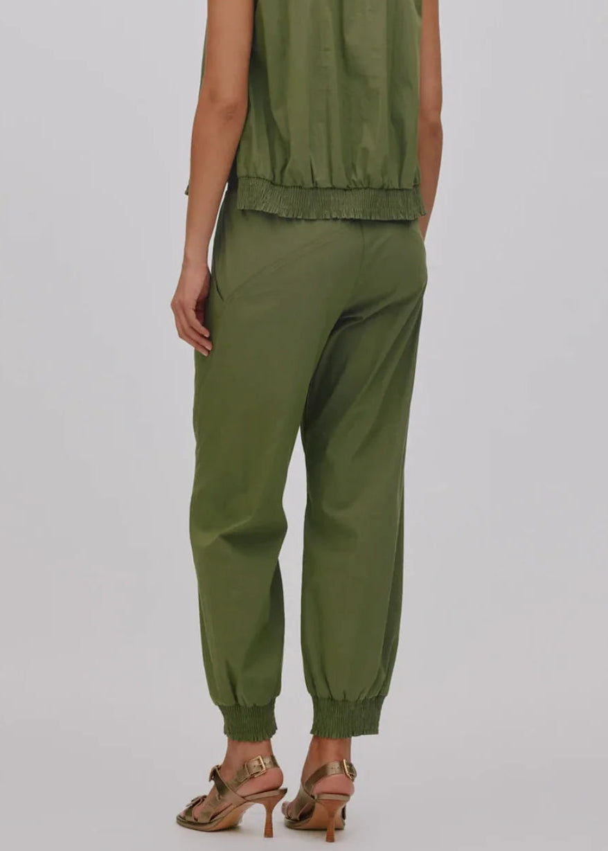 A woman shown from the back wearing an olive green sleeveless top and matching jogger pants, paired with brown heeled sandals, against a light gray studio background.