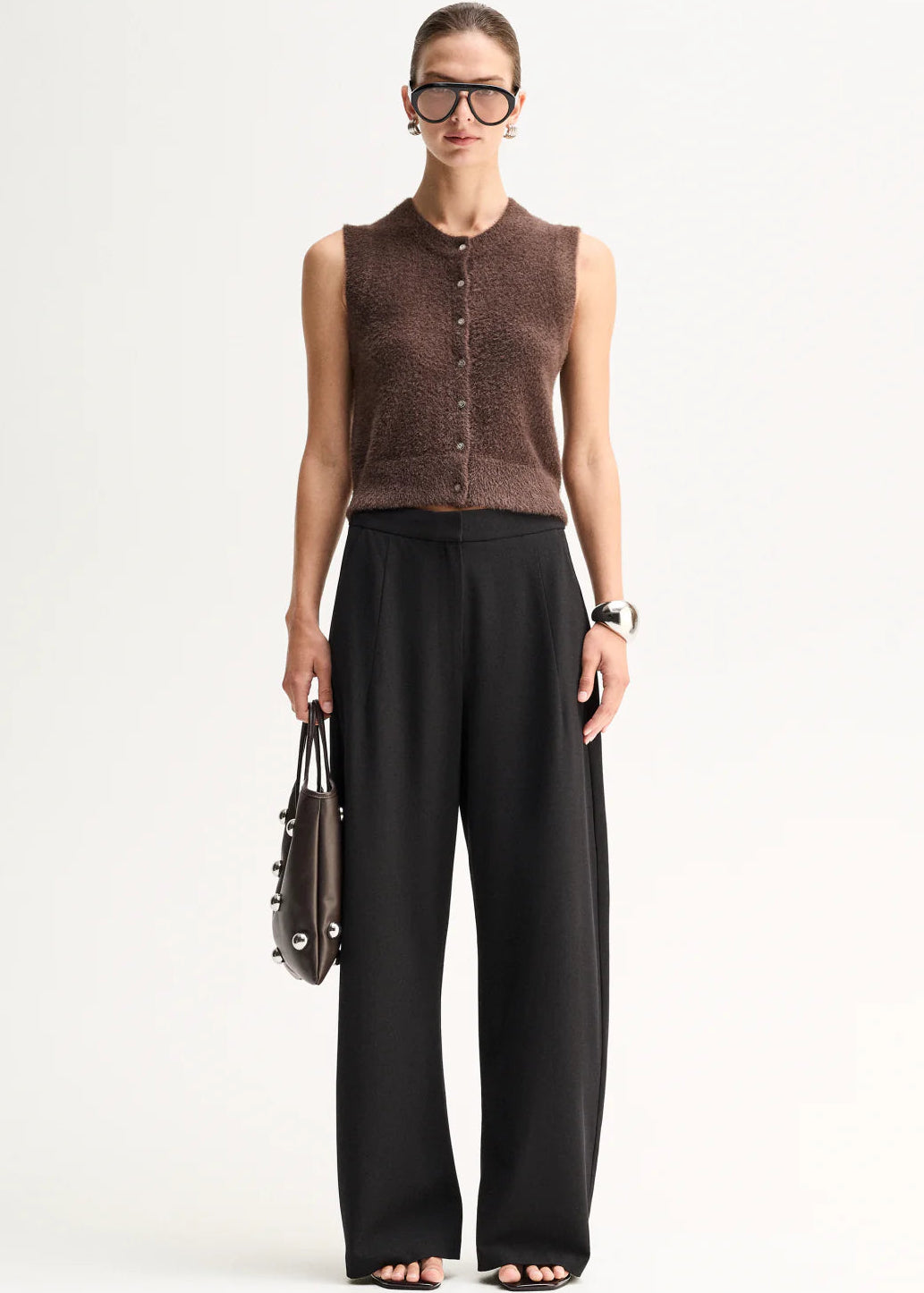 A model stands against a plain light background wearing a brown sleeveless button-front knit top,