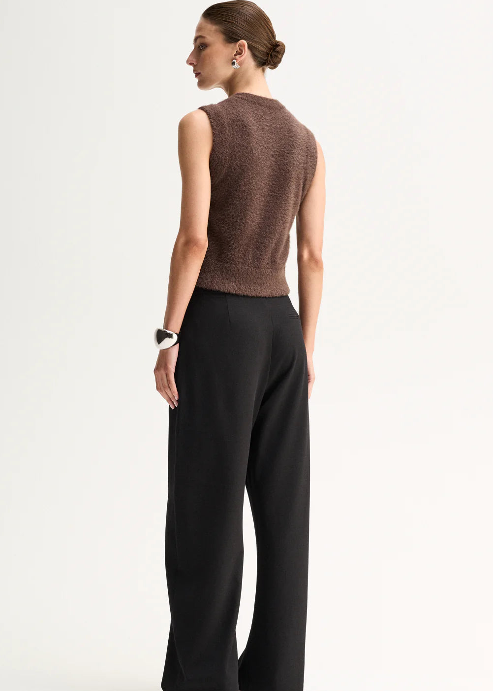 A model stands against a plain light background wearing a brown sleeveless button-front knit top,