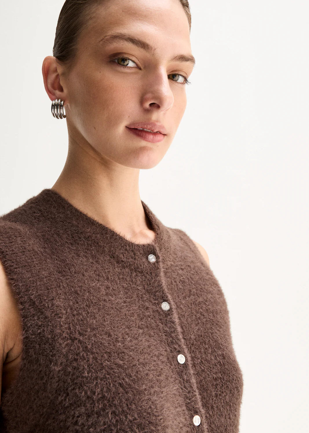 A model stands against a plain light background wearing a brown sleeveless button-front knit top,