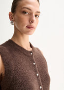 A model stands against a plain light background wearing a brown sleeveless button-front knit top,