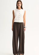 A woman stands against a plain light background wearing a sleeveless white knit vest with a button front and dark brown wide-leg trousers.