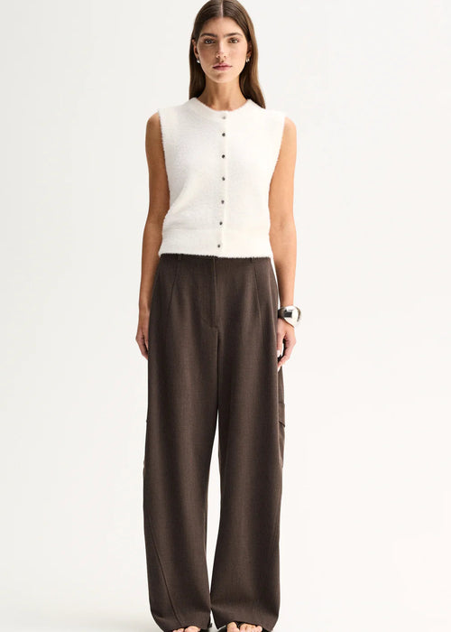 A woman stands against a plain light background wearing a sleeveless white knit vest with a button front and dark brown wide-leg trousers.