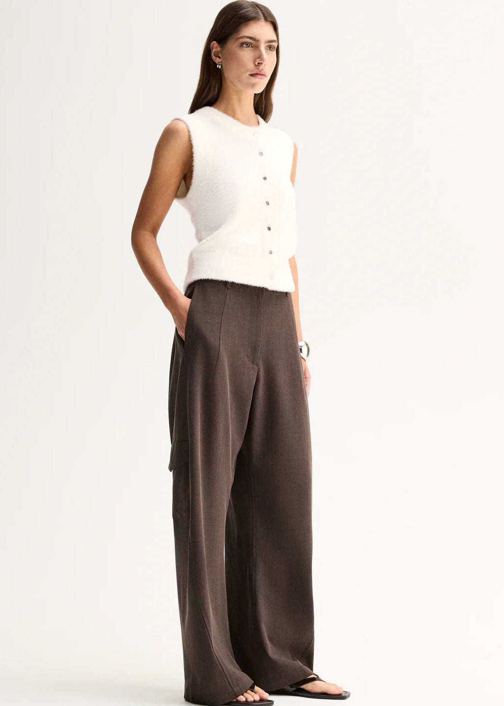 A woman stands against a plain light background wearing a sleeveless white knit vest with a button front and dark brown wide-leg trousers.