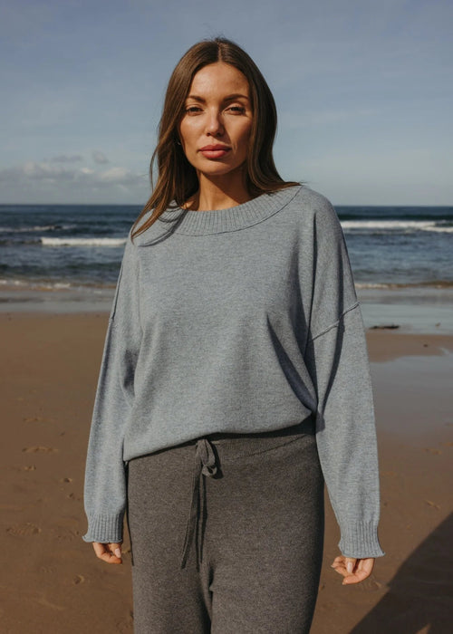 Woman wearing a gray sweater and dark pants standing on a beach with ocean waves in the background.