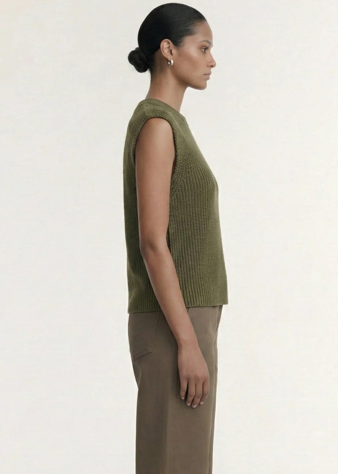 A model wearing an olive green ribbed sleeveless knit top and brown wide-leg trousers, standing against a plain off-white studio background.