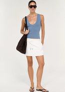 A woman modelling a light blue knit tank top and white drawstring mini skirt, carrying a large black woven tote bag, against a plain light grey studio background.