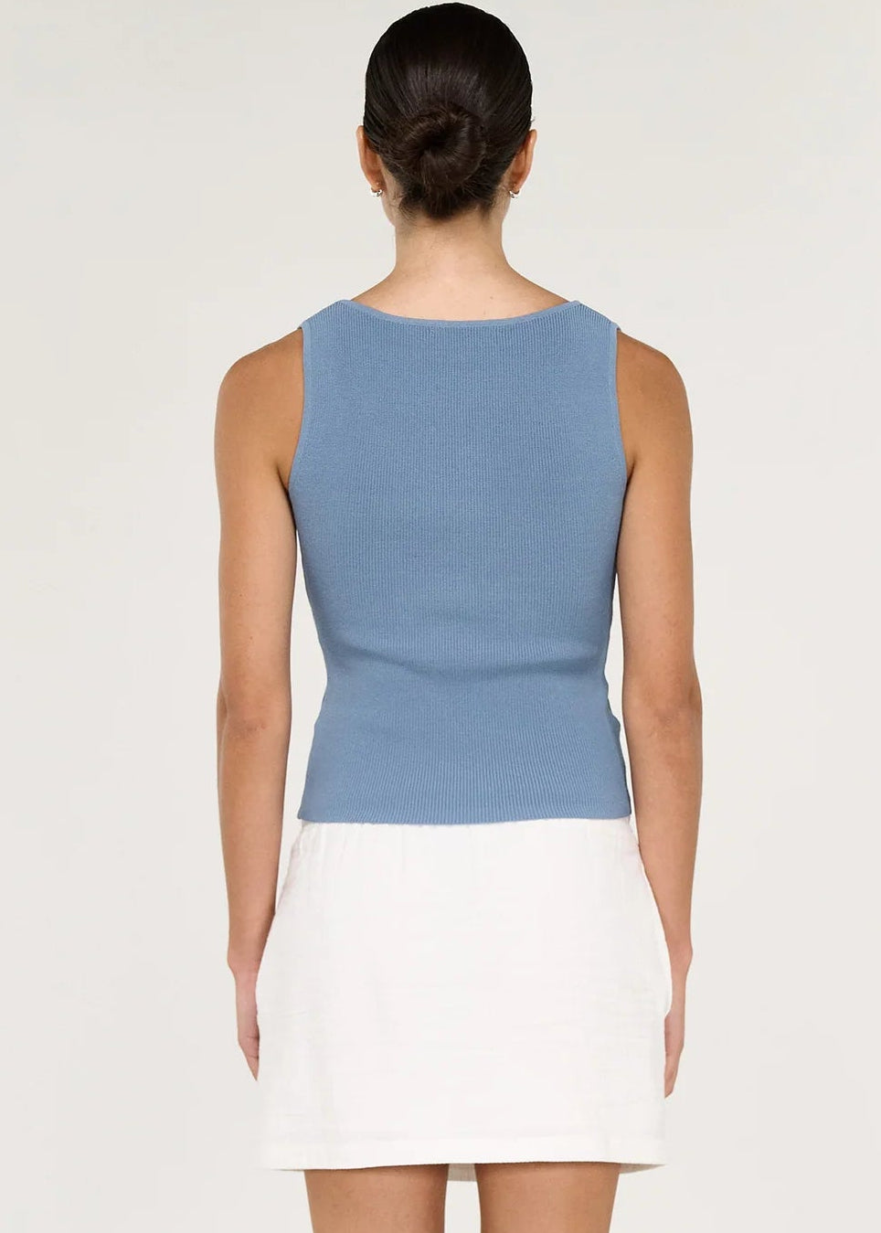 A woman viewed from the back wearing a sleeveless knit top with a light blue upper section and a white skirted lower section, paired with black flat sandals against a plain light background.