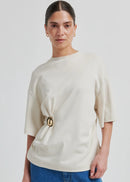 Woman wearing a cream-colored top with a brooch and blue jeans on a white background